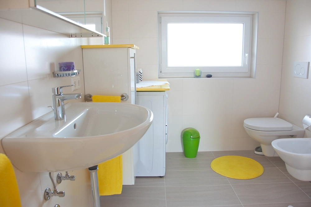 Ferienwohnung Greiler – Innenansicht, kleines Badezimmer mit Waschbecken und Badezimmermöbel; in weiß und gelb gehalten, helle, freundliche Lichtstimmung.